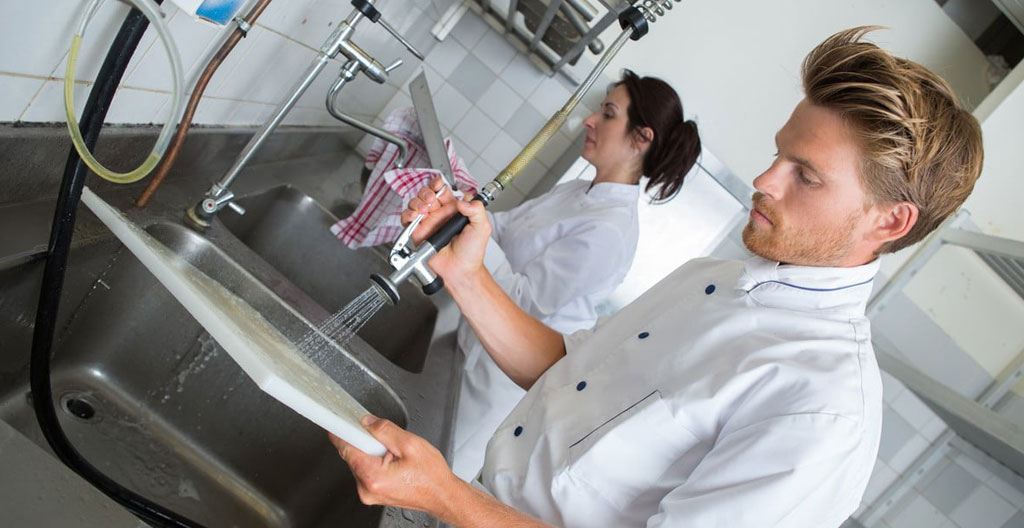Un employé vu de dos charge des casiers devant un lave-vaisselle à capot dans une zone de plonge en inox.